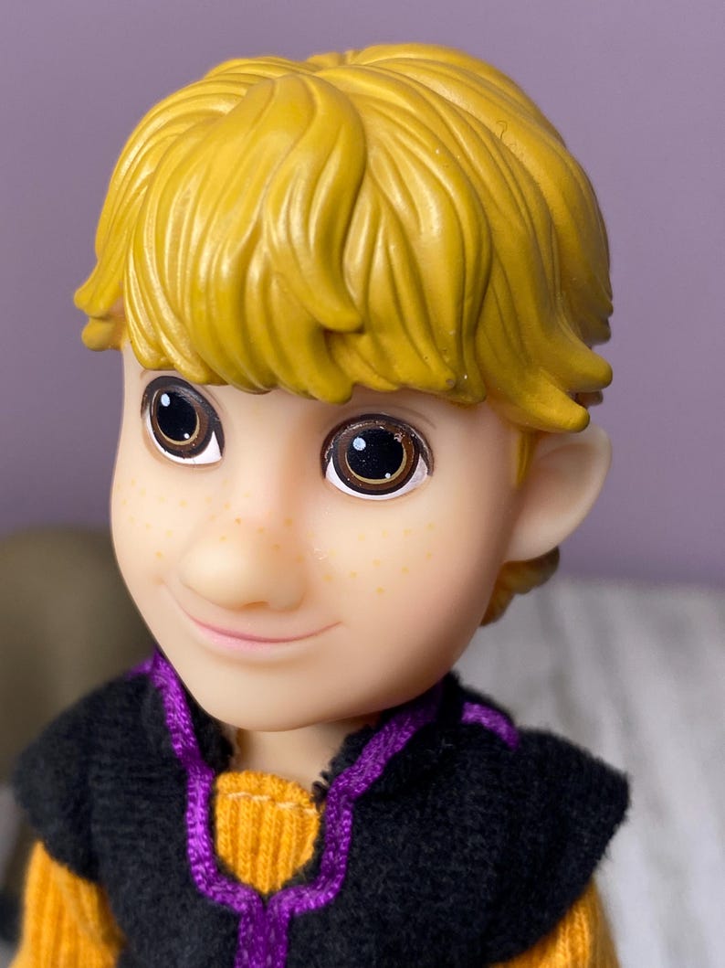 K&ouml;nnte beinhalten: Nahaufnahme eines Puppengesichts mit gelbem Haar und braunen Augen. Die Puppe hat einen hellen Teint mit Sommersprossen und tr&auml;gt eine schwarze und lila Weste &uuml;ber einem orangefarbenen Pullover. Die Puppe l&auml;chelt.