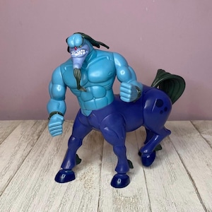 May include: A blue and purple centaur action figure. The figure has a muscular upper body with a blue complexion and a horse-like lower body in a darker shade of blue. The figure has a stern expression and is posed with its fists clenched.