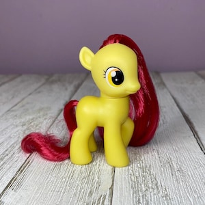 May include: A yellow toy pony with a red mane and tail. The pony has large eyes with black pupils and yellow and white accents. The toy is standing on a wooden surface.
