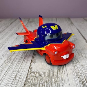 May include: A red toy car with blue and yellow wings and a blue helmet. The car has a smiling face and the letter "M" on the helmet. The toy car is on a light wood surface.