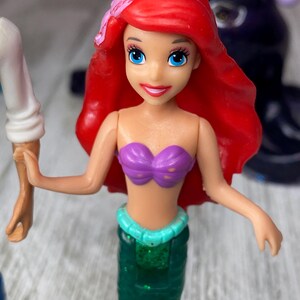 May include: Ariel doll with red hair, blue eyes, and a purple seashell top. The doll has a green mermaid tail with a turquoise waist band. A pink flower is in her hair. Another figure is in the background.