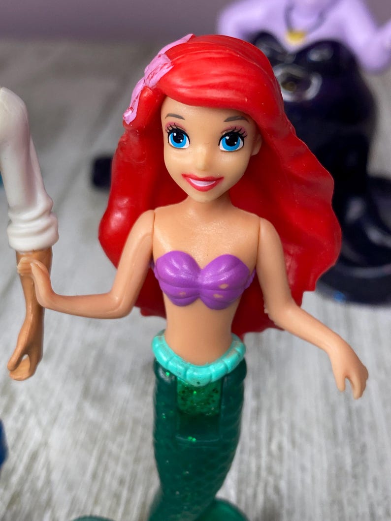 May include: Ariel doll with red hair, blue eyes, and a purple seashell top. The doll has a green mermaid tail with a turquoise waist band. A pink flower is in her hair. Another figure is in the background.