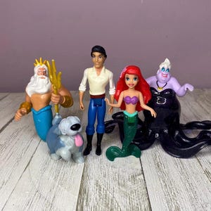 May include: A collection of Disney figurines featuring characters from The Little Mermaid. The set includes Ariel, Prince Eric, Ursula, King Triton, and a dog. The figurines are brightly colored and made of plastic.
