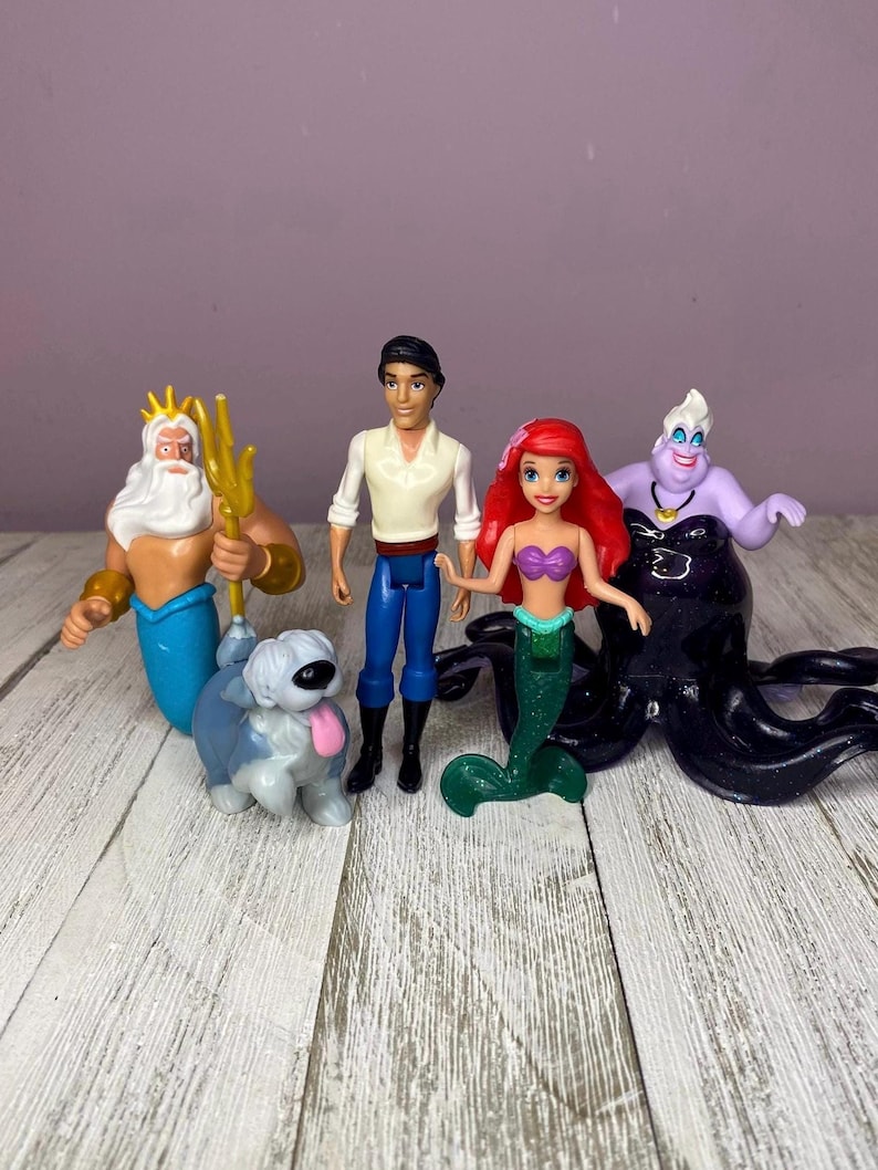 May include: A collection of Disney figurines featuring characters from The Little Mermaid. The set includes Ariel, Prince Eric, Ursula, King Triton, and a dog. The figurines are brightly colored and made of plastic.