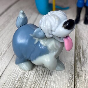 May include: A small, blue and white toy dog figurine with a pink tongue sticking out. The dog has a fluffy white head and a black nose. Other toy figures are visible in the background.