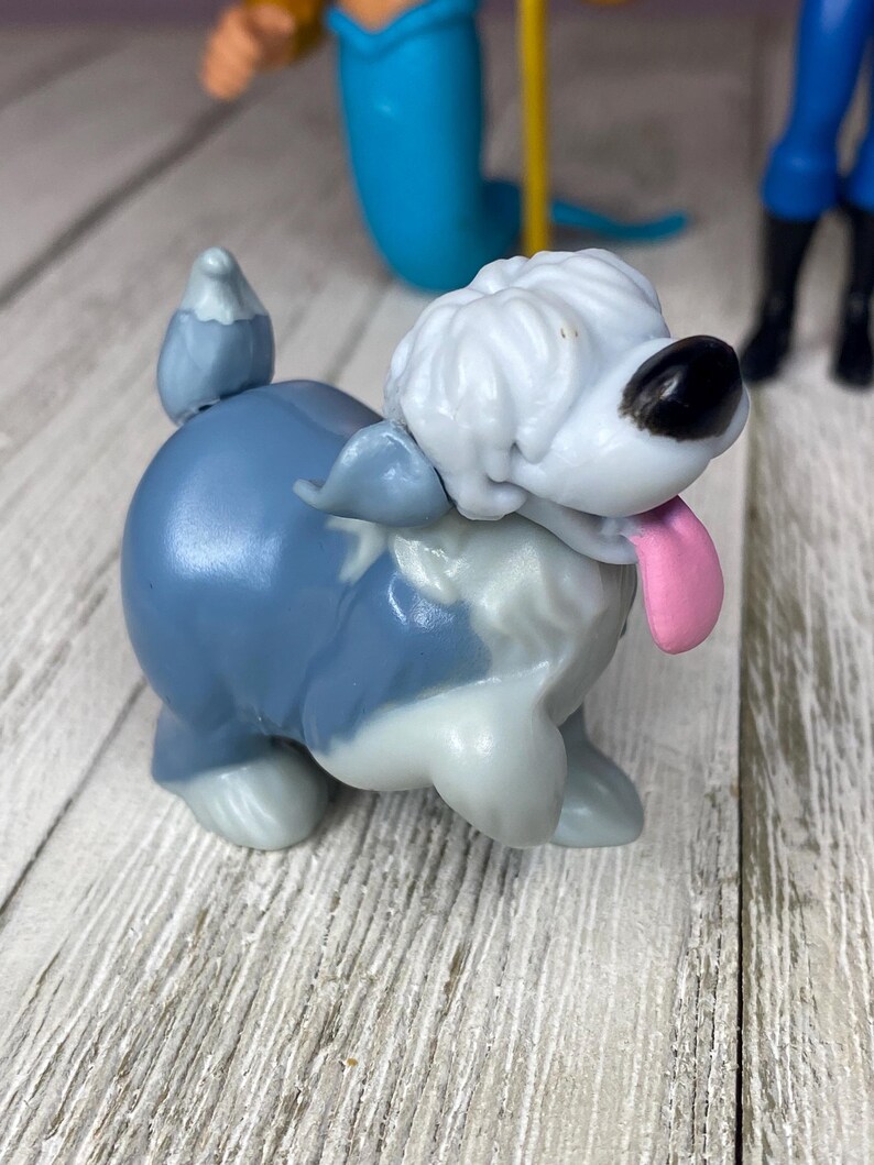 May include: A small, blue and white toy dog figurine with a pink tongue sticking out. The dog has a fluffy white head and a black nose. Other toy figures are visible in the background.