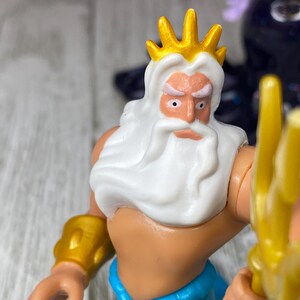 May include: A plastic figurine of King Triton, featuring a gold crown, white beard, and blue pants. He holds a gold trident and wears gold arm cuffs. The figure has a stern expression.