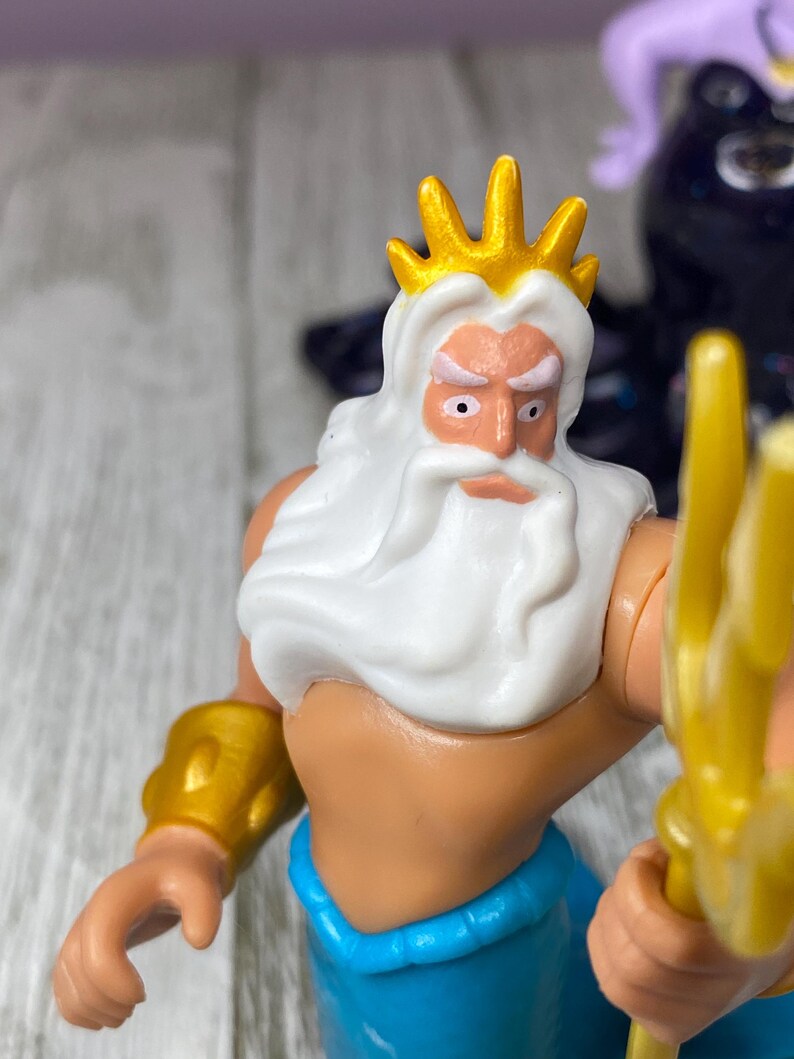 May include: A plastic figurine of King Triton, featuring a gold crown, white beard, and blue pants. He holds a gold trident and wears gold arm cuffs. The figure has a stern expression.