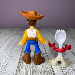 May include: A Woody action figure from the Toy Story franchise, wearing a brown cowboy hat, yellow shirt, white vest, blue pants, and brown boots. A white and red spork toy is next to the figure.