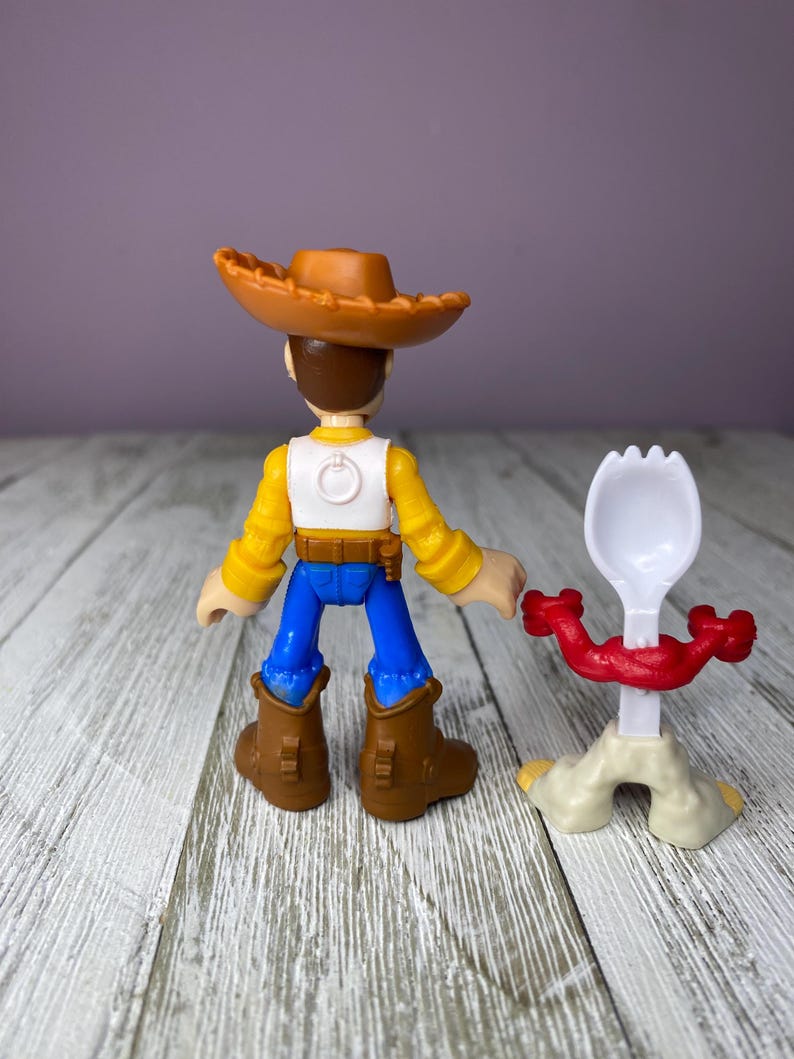 May include: A Woody action figure from the Toy Story franchise, wearing a brown cowboy hat, yellow shirt, white vest, blue pants, and brown boots. A white and red spork toy is next to the figure.