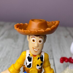 May include: A Woody action figure from the Toy Story franchise. The figure has a brown cowboy hat, a yellow shirt, a black and white vest, blue pants, and a sheriff's badge. The figure is made of plastic and is a collectible item.