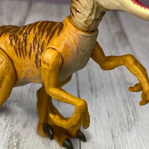 May include: A detailed close-up of a yellow and brown toy dinosaur, likely a Velociraptor. The toy has articulated joints and features brown stripes on its back. The dinosaur's mouth is open, revealing teeth and a red interior.