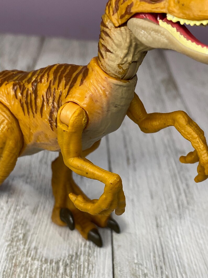 May include: A detailed close-up of a yellow and brown toy dinosaur, likely a Velociraptor. The toy has articulated joints and features brown stripes on its back. The dinosaur's mouth is open, revealing teeth and a red interior.