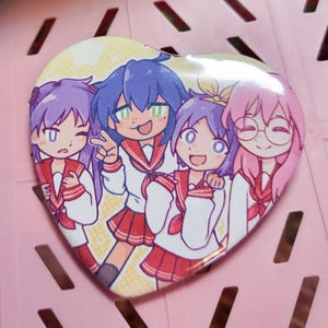 May include: Heart-shaped button featuring an anime-style illustration of four girls in school uniforms. The girls have different hair colors: purple, blue, and pink. They are smiling and posing, with one making a peace sign.