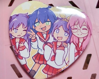 May include: Heart-shaped button featuring an anime-style illustration of four girls in school uniforms. The girls have different hair colors: purple, blue, and pink. They are smiling and posing, with one making a peace sign.