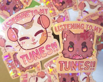 Könnte beinhalten: Eine Sammlung holografischer Aufkleber mit Cartoon-Katzen. Die Aufkleber zeigen den Text "LISTENING TO MY TUNES!!" in Rosa und Orange. Die Katzen sind in verschiedenen Farben gehalten, darunter Weiß, Braun und Rosa, und einige tragen Kopfhörer oder Ohrhörer.
