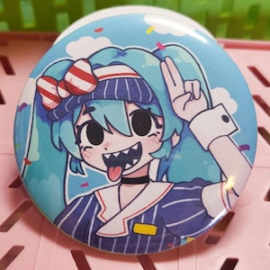 May include: A round, blue button featuring an anime-style character with turquoise hair, a striped bow, and a navy and white striped outfit. The character is sticking out their tongue and making a peace sign with their fingers.