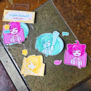 May include: A sticker pack featuring anime-style illustrations. The stickers include characters in pink, yellow, and teal, with speech bubbles and the text "triple baka!!! sticker pack". The stickers are on a clear backing.