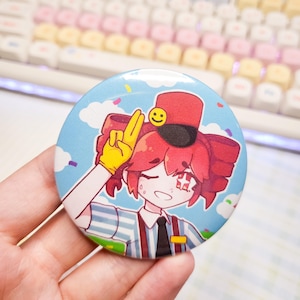 May include: A circular pinback button featuring an anime-style character with red hair, wearing a red hat with a yellow smiley face, a blue and white striped shirt, and a yellow glove. The character is making a peace sign against a blue sky background.