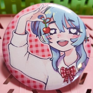 May include: A round button with a cartoon illustration of a girl with blue hair holding a cherry. The button has a pink and red checkered background. The girl has heart-shaped eyes and a red bow.