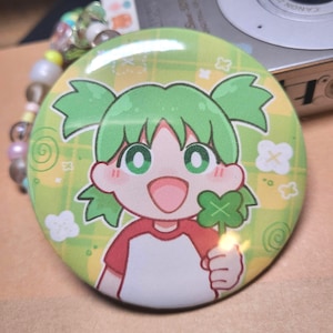 May include: A circular badge with a cartoon girl with green hair and eyes, wearing a red and white shirt, holding a four-leaf clover. The badge has a green and yellow plaid background with white clover accents.