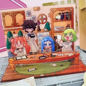 May include: A multi-layered acrylic standee featuring four illustrated characters in a room with a wooden table, bookshelves, and cabinets. The characters have distinct hair colors including red, dark brown, blue, and light green, and are depicted in a cartoon style with various small items like books, papers, and ink pots scattered on the table.
