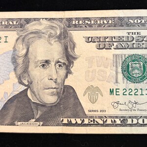 May include: A twenty-dollar bill featuring a portrait of Andrew Jackson. The bill displays the text "The United States of America" and "Twenty Dollars". The serial number is ME 22213222 I.