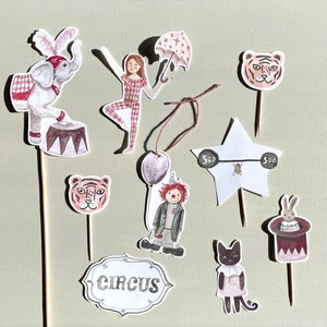 Könnte beinhalten: Ein Set mit 10 bunten Cupcake-Toppern zum Thema Zirkus. Die Topper zeigen einen rosa Elefanten, einen rosa Tiger, einen rosa-weißen Clown, eine rosa-weiße Katze, einen rosa-weißen Hasen, einen rosa-weißen Stern, einen rosa-weißen Regenschirm, ein rosa-weißes Zirkus-Schild, eine rosa-weiße Hantel und einen rosa-weißen Hut.