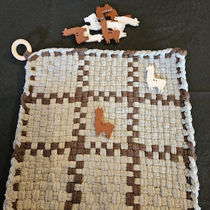 May include: A woven gray and brown square pot holder with a wooden ring and llama-shaped embellishments. The pot holder has a woven pattern with brown squares and a brown border. Several llama-shaped wooden cutouts are scattered around the pot holder.