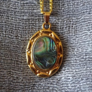 Könnte beinhalten: Eine goldfarbene Halskette mit einem ovalen Anhänger. Der Anhänger zeigt eine schillernde, mehrfarbige Abalone-Muschel, die in einen dekorativen Goldrahmen gefasst ist. Die Kette ist eine zarte Goldkette, geeignet für den täglichen Gebrauch.