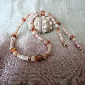 Peut inclure: Un collier de perles rose et blanc avec des accents floraux et des accents dorés. Le collier est sur un fond de tissu blanc et rose.