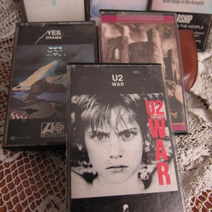以下が含まれることがあります： 5 本のカセットテープのコレクション。U2 の「War」カセットテープは、前面に若い人の白黒写真が印刷されています。Yes の「Drama」カセットテープは、前面に青と白の風景が印刷されています。もう 1 本のカセットテープは、前面に城の写真が印刷されています。カセットテープは、白いレースの布の上に置かれています。