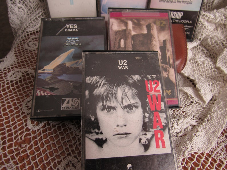 以下が含まれることがあります： 5 本のカセットテープのコレクション。U2 の「War」カセットテープは、前面に若い人の白黒写真が印刷されています。Yes の「Drama」カセットテープは、前面に青と白の風景が印刷されています。もう 1 本のカセットテープは、前面に城の写真が印刷されています。カセットテープは、白いレースの布の上に置かれています。