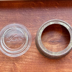 May include: A clear glass Mason jar lid with the word "Mason" embossed on it, next to a rusty metal jar ring. The lid is round with concentric circles. The jar ring is a weathered bronze color with a green hue.
