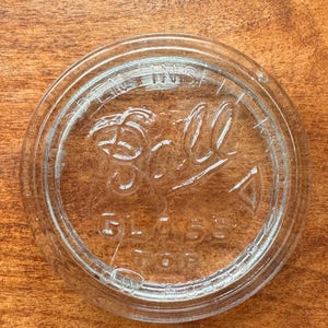 May include: A circular clear glass lid featuring embossed lettering that reads Ball Glass Top and Open Inside. The lid has a raised outer rim and a recessed center section, designed to fit onto a jar for storage or preservation purposes.