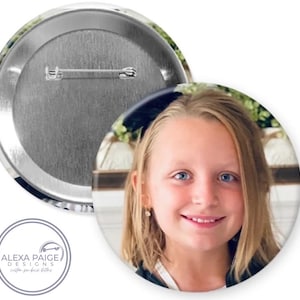 May include: A round button with a photo of a young girl with blonde hair and blue eyes. The girl is smiling and looking at the camera. The button has a silver back with a safety pin.