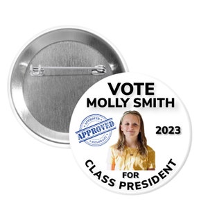 May include: White button with a black border and text that reads "Vote Molly Smith for Class President 2023". A blue stamp with the word "Approved" is in the upper left corner. A photo of a young girl is in the center of the button.