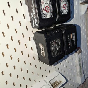 May include: Black Makita 18V 3.0Ah and 9.0Ah Li-ion tool batteries mounted on a white pegboard. The pegboard also holds small white containers filled with screws and other hardware. The batteries are labeled with safety information.