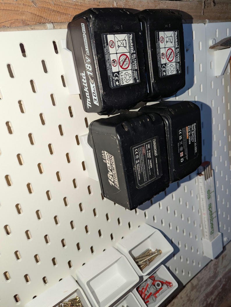 May include: Black Makita 18V 3.0Ah and 9.0Ah Li-ion tool batteries mounted on a white pegboard. The pegboard also holds small white containers filled with screws and other hardware. The batteries are labeled with safety information.