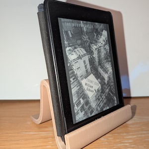 Könnte beinhalten: Ein schwarzer E-Reader mit Buchcover wird auf einem hellbraunen, gebogenen Ständer präsentiert. Der Bildschirm des E-Readers zeigt das Buchcover von "Count Zero" von William Gibson. Der Ständer besteht aus hellbraunem Material und steht auf einer Holzoberfläche.