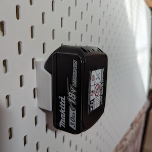 May include: A black Makita 18V 3.0Ah Lithium-Ion battery pack mounted on a white pegboard. The battery is secured by a white plastic holder. The pegboard has evenly spaced rectangular holes.