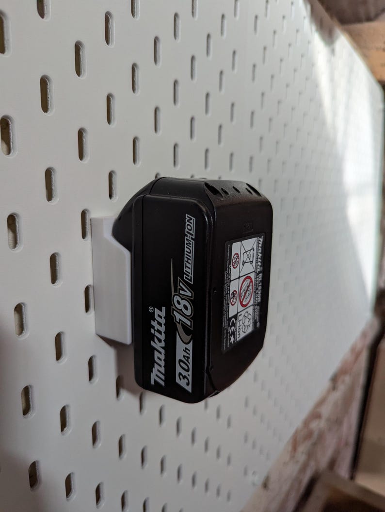 May include: A black Makita 18V 3.0Ah Lithium-Ion battery pack mounted on a white pegboard. The battery is secured by a white plastic holder. The pegboard has evenly spaced rectangular holes.