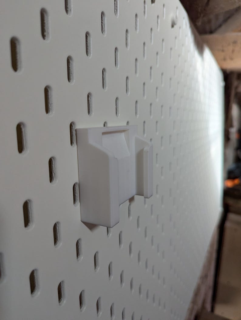 May include: A white pegboard with a white plastic holder attached. The pegboard has evenly spaced, vertical slots. The holder is designed to fit into the slots, providing a mounting point for tools or other items.