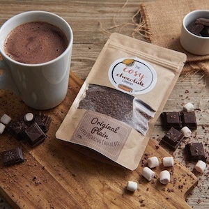 Puede incluir: Una bolsa de papel marrón con el texto "The Cosy Chocolate Company" y "Original Plain Hot Drinking Chocolate" impreso en ella. La bolsa está llena de virutas de chocolate y está sentada sobre una mesa de madera junto a una taza de chocolate caliente, malvaviscos y cuadrados de chocolate.