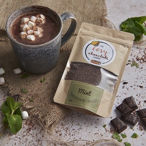 Puede incluir: Una bolsa de papel marrón con una ventana transparente que muestra un polvo marrón oscuro en su interior. La bolsa tiene una etiqueta blanca con el texto "The Cosy Chocolate Company" y una etiqueta más pequeña que dice "Chocolate para beber con sabor a menta". También hay una taza de chocolate caliente con malvaviscos en la parte superior, algunos malvaviscos esparcidos sobre una superficie de arpillera y virutas de chocolate sobre una superficie gris claro.
