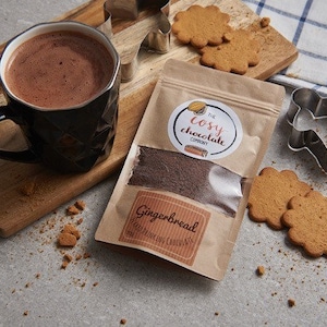 Puede incluir: Una bolsa de papel marrón con una ventana transparente que muestra un polvo marrón oscuro en su interior. La bolsa tiene una etiqueta blanca con el texto "The Cosy Chocolate Company" y "Gingerbread" impreso en ella. En el fondo se ven galletas de jengibre y una taza de chocolate caliente.