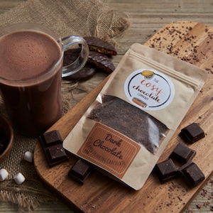 Puede incluir: Una bolsa de papel marrón llena de polvo de cacao de chocolate negro. La bolsa tiene una etiqueta que dice "The Cosy Chocolate Company" y "Dark Chocolate". También hay cuadrados de chocolate y una taza de chocolate caliente en una tabla de cortar de madera.