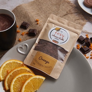 Puede incluir: Una bolsa de papel marrón con una etiqueta blanca que dice "The Cosy Chocolate Company" y "Chocolate para beber con sabor a naranja". La bolsa está sobre un plato gris con rodajas de naranja y cuadrados de chocolate.