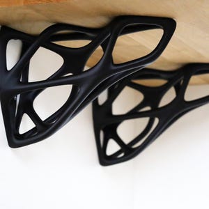 May include: Two black, decorative shelf brackets with an open, geometric design. The brackets are mounted beneath a light-colored wooden shelf, creating a modern, minimalist aesthetic. The brackets are made of a solid material.