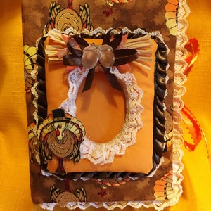 May include: A handmade Thanksgiving-themed photo album. The cover features a brown fabric with turkey illustrations, accented with white lace trim and a decorative frame. Embellishments include a straw bow, brown ribbon, and acorn accents. A festive and decorative item.